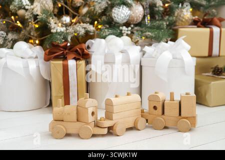 Wooden toy train with wagons placed on white floor in front of wrapped Christmas gift boxes and decorated tree with ornaments and shining lights. Atmo Stock Photo