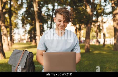 Cheerful student guy with laptop computer doing homework or attending online lecture, sitting on bench outdoors Stock Photo