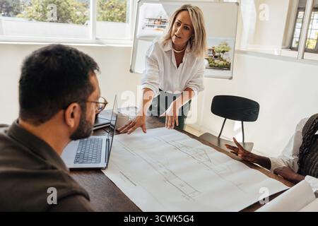 Three architects collaborating around a table reviewing building plans in a bright office. The discussion includes two males and a female, showcasing Stock Photo