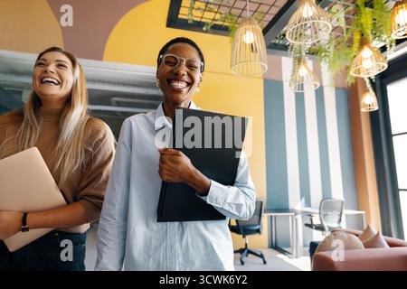 Two young women stand in a modern office, smiling while holding their work essentials. Stock Photo