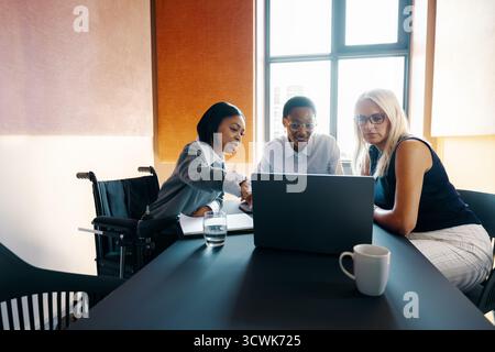 Three women of diverse backgrounds discussing work at a laptop in a modern office. One woman is a wheelchair user, highlighting inclusivity and teamwo Stock Photo