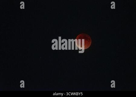 A wide, atmospheric view of the total lunar eclipse, showing the solitary Blood Moon as a small, deep red orb in the vast, dark emptiness of a star-du Stock Photo