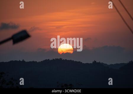 The glowing orange sun sets behind rolling hills, with the dark silhouettes of a streetlight and power lines in the foreground. A scenic view of dusk Stock Photo