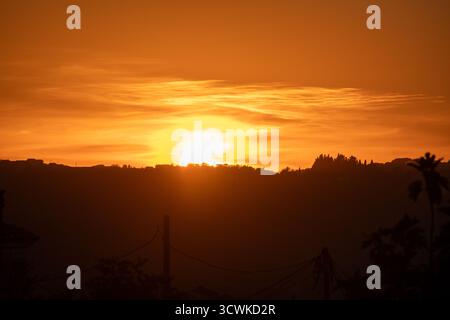 The sun bursts with intense golden light as it sets behind silhouetted hills in Greece. A powerful and vibrant sunset scene, painting the entire sky a Stock Photo