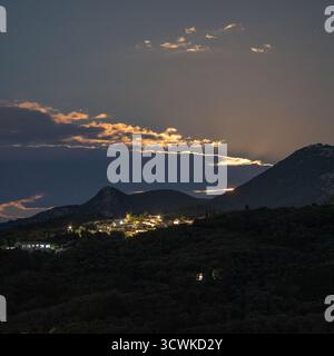The moon rises behind mountains, casting a golden glow on the clouds above a tranquil, illuminated village in the Greek countryside at night. A serene Stock Photo