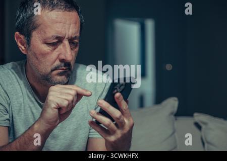 Tired mature man typing text message on smartphone in dark living room interior, using mobile phone with index finger, low key portrait in casual home Stock Photo