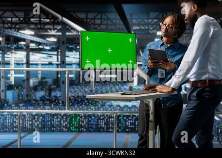 African american engineer uses copy space on computer monitor in server hub, analyzing software systems data while he stands on industrial platform. Engineering AI tech configurations. Stock Photo