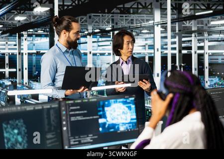 Multiethnic coworkers coding in diverse data center, updating AI automation scripts. Multiracial server room colleagues doing brainstorming, programming artificial intelligence algorithms Stock Photo