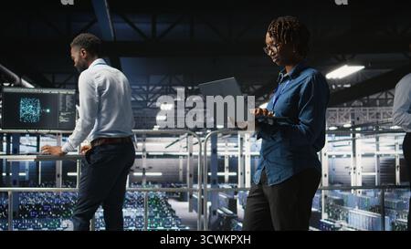 System administrators on data center industrial platform analyzing performance outputs on laptop. Server hub teamworking colleagues finetuning server rig configurations using notebook, Stock Photo