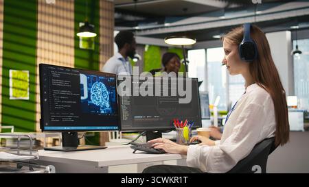 Young female programmer in office reviewing AI system metrics, listening music, monitoring performance dashboards. Junior dev wearing headphones tuning neural networks using tools on PC, camera A Stock Photo