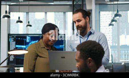 Teamworking programmers in office reviewing AI system metrics, monitoring performance dashboards. Tech company coworkers tuning neural networks using tools on laptop, camera A Stock Photo