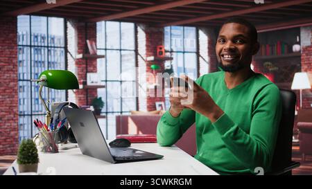 Man taking break from work to check his social media apps on mobile phone, having fun relaxing at home. Young guy in loft pausing tasks to use social network application on phone. Camera B. Stock Photo
