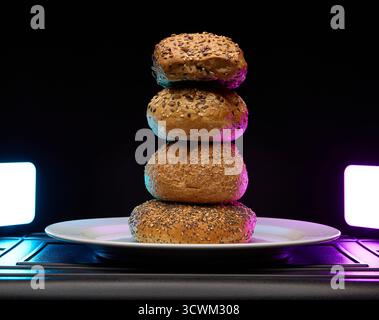 Four assorted bagels stacked vertically on a plate, illuminated by modern purple and cyan studio light. Contemporary bakery still life for advertising Stock Photo