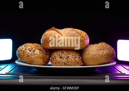 Group of assorted bread buns and bagels piled on a plate under soft purple and blue light. Modern still life bakery image for commercial use or food a Stock Photo
