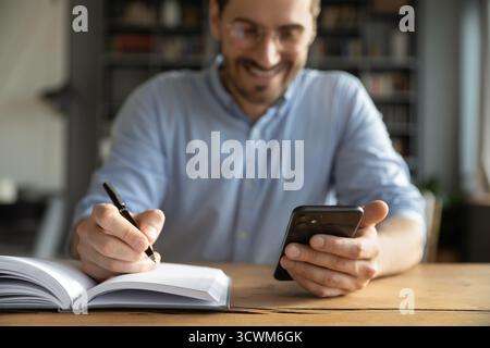 Closeup young businessman holding smartphone writing with pen at desk Stock Photo