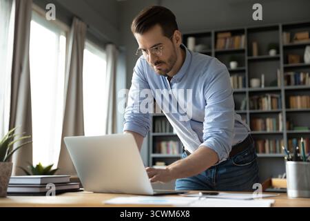 Concentrated young businessman standing by workplace desk writing on laptop Stock Photo
