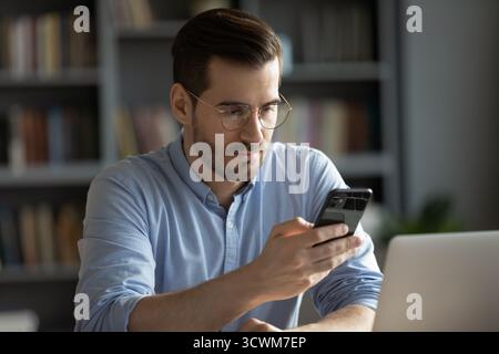 Focused young businessman sitting at desk by notebook using smartphone Stock Photo