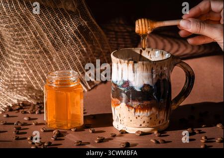 A honey dipper dripping honey into a ceramic coffee mug next to an open jar of honey, coffee beans spilled in foreground, brown background Stock Photo