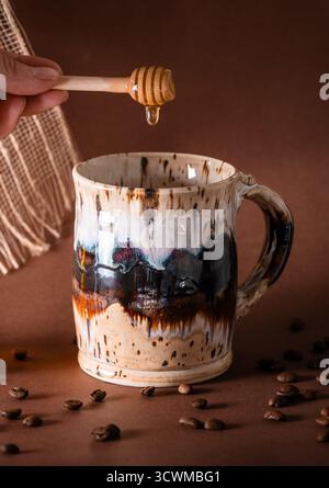 A honey dipper dripping honey into a ceramic coffee, coffee beans spilled in foreground, brown background Stock Photo