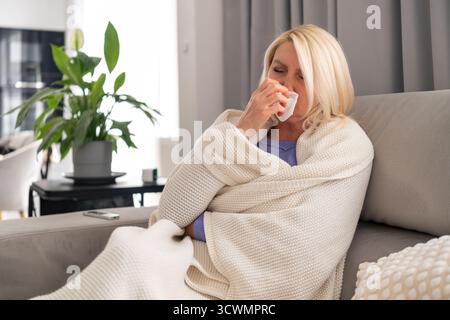 Middle aged woman wrapped in plaid sitting on couch and blowing her nose into a tissue. Concept of flu, covid, home rest, and self care during illness. Stock Photo