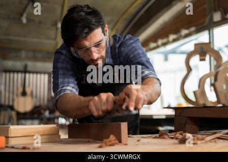 Professional carpenter working with wood planer on wooden surface in workshop, wearing protective glasses and apron, demonstrating focus, craftsmanshi Stock Photo
