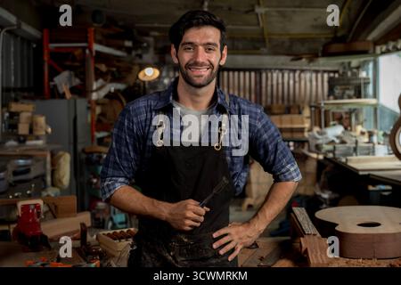 Confident male carpenter smiling in workshop while holding chisel, wearing safety apron and casual shirt, representing craftsmanship, small business o Stock Photo