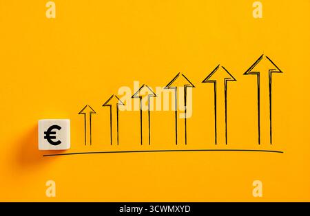 Wooden block with euro currency symbol and increasing upward arrows drawn on yellow background representing financial growth and positive investment Stock Photo