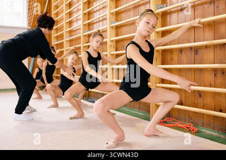 Group of Caucasian girls practicing ballet exercises at barre under guidance of middle aged Caucasian woman instructor, girls performing plie positions in dance studio, focused expressions Stock Photo