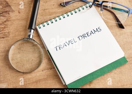 A notepad with the words TRAVEL DREAMS sits on a wood surface beside a magnifying glass and glasses. This suggests someone is planning their next trip Stock Photo