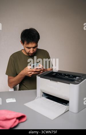Focused man using smartphone to read instructions while repairing laser printer, with cleaning supplies and tools nearby on table, on isolated Stock Photo