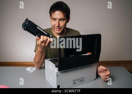 Cheerful computer technician replacing toner cartridge in laser printer during maintenance service, holding new cartridge and looking away Stock Photo