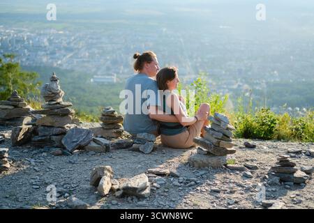 Young couple sitting back view embracing while admiring breathtaking cityscape from mountain top, with stone cairns in foreground. Perfect for travel and love concepts. Stock Photo