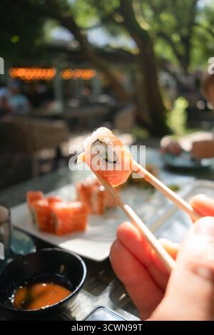 Appetizing sushi roll filled with fresh salmon and crisp cucumber, captured mid-air with chopsticks against restaurant backdrop. Stock Photo