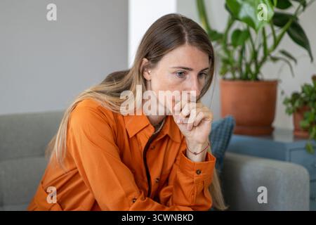 Worried middle-aged woman touching her chin deeply absorbed in thought sitting alone on sofa Stock Photo