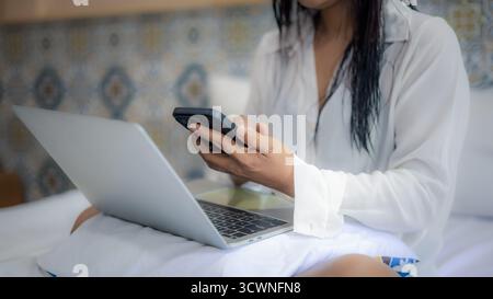 Close-up of woman using smartphone and laptop while working remotely in bed. Modern digital nomad lifestyle, freelance or work from anywhere concept. Stock Photo
