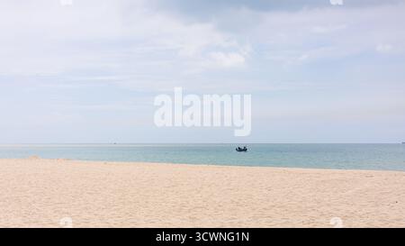 Minimal seascape with golden sand beach, calm blue ocean, and small fishing boat under cloudy sky, peaceful travel and relaxation background. Stock Photo