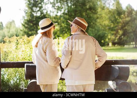 Two stylish mature women standing back, wearing light beige jackets and summer hats, enjoying sunny day in the park. Relaxed conversation, friendship, Stock Photo