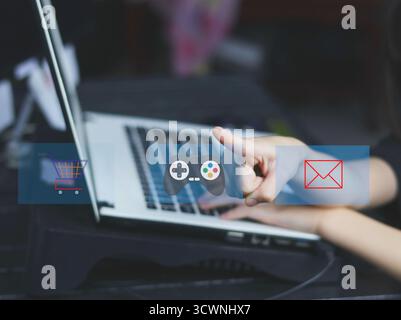 An image of a person's hand pointing at a floating game controller icon above a laptop keyboard. Other icons include a shopping cart and an envelope. Stock Photo
