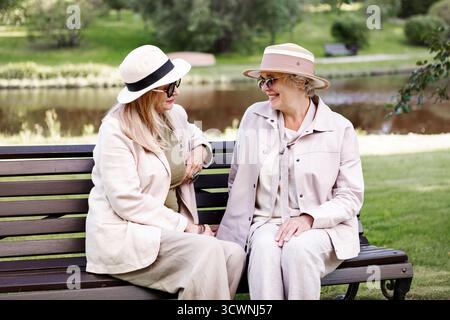 Two happy senior women sitting on park bench and talking happily near pond, enjoying friendship and leisure outdoors, stylish elderly friends in light Stock Photo