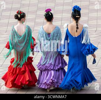 Sevilla, Spain - 23 Apr 23: Three women in traditional flamenco dresses walking away, showcasing vibrant colors and intricate designs, with floral hai Stock Photo