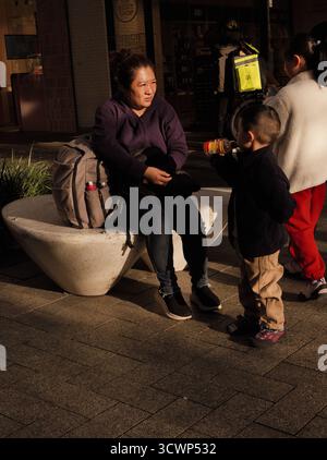 Mother and child resting during the golden hour on a city street; deep shadows and warm sunlight create a moody, tender, and reflective scene. Stock Photo