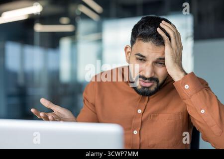 Stressed young male employee experiencing headache and frustration, holding hand on head while looking at laptop screen in modern office, indicating work burnout or data loss Stock Photo
