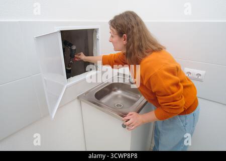 Woman plumber turning off water valve under sink. Female hand adjusting pipe in utility closet. Home repair emergency concept. Stock Photo