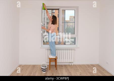 Woman cleaning apartment window on stool with rag, cleaning house or apartment as concept of housework and home chores for new building space. Stock Photo