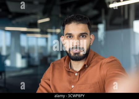 Young indian man in brown shirt confidently taking a selfie in a modern office, creating professional social media content and showcasing entrepreneur style and smart-casual confidence Stock Photo