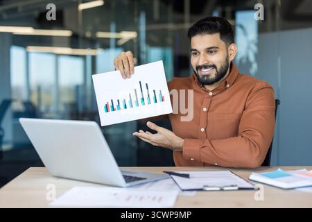 Young businessman presenting data analysis on a bar chart during an online video conference, collaborating with colleagues while working remotely from a modern office Stock Photo