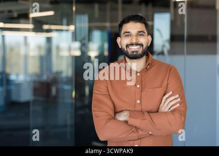 Young indian businessman with a beard and confident smile posing with arms crossed, standing in a blurred modern office environment, representing professionalism, success, and corporate leadership Stock Photo
