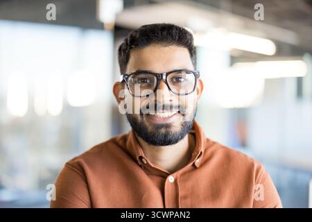 Young indian man with a beard and glasses smiling confidently at the camera, portraying a successful and approachable professional in a modern office environment Stock Photo