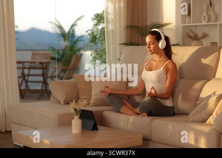 Yogi doing online guided yoga class sitting on a couch at home at sunset Stock Photo
