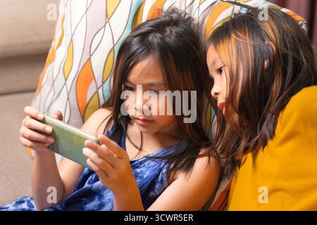 Two Asian girls sitting on sofa watching smartphone screen together at home, representing childhood, learning, technology, and digital lifestyle in mo Stock Photo